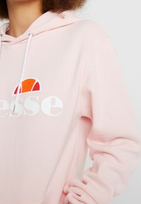 Sudadera con capucha rosa con un logo blanco, que presenta un diseño circular colorido en el pecho y cuerdas blancas en la capucha.