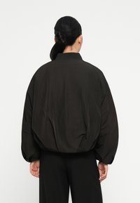 Zwarte, oversized bomberjack met een matte afwerking, geribbelde kraag en volumineuze mouwen, gezien van achteren tegen een witte achtergrond.