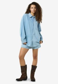 Chemise en denim oversize bleu clair avec fermeture à boutons et poche, assortie à un short rayé, portée avec des bottes de cowboy marron.