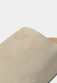 Μπεζ σουέντ σανδάλι slide με ανάγλυφο λογότυπο trefoil της Adidas στο φαρδύ λουράκι, σε κοντινή λήψη με φόντο λευκό.