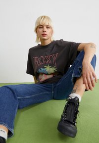 Čierne grafické tričko s nápisom "ROXY" a dizajnom palmovej tree, v kombinácii s modrými džínsmi a čiernymi šnurovacími teniskami na zelenej ploche.