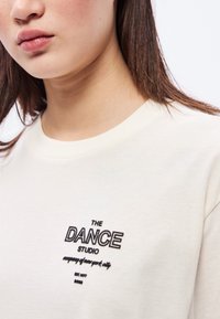 Svetlokrémové tričko s okrúhlym výstrihom a čiernym vyšitým textom: "THE DANCE STUDIO company of new york city EST. 1977."