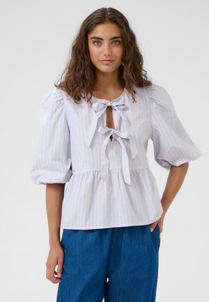 Jonge vrouw met krullend bruin haar, gekleed in een lichtblauwe en beige gestreepte blouse met pofmouwen en strikjes aan de voorkant, gecombineerd met een blauwe broek.