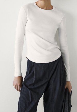 Personne portant un haut blanc à manches longues et un pantalon ample à fines rayures sombres, tenant un tissu texturé foncé dans une main.