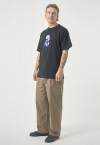 Schwarzes Baumwoll-T-Shirt mit einem lila grafischen Design auf der Brust, kombiniert mit beigen weit geschnittenen Hosen und grünen Turnschuhen.