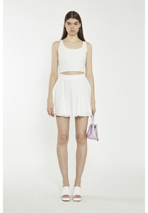 Ung kvinde iført en hvid crop top og nederdel med hulmønster, der holder en lille skinnende pink håndtaske, stående foran en ensfarvet hvid baggrund.