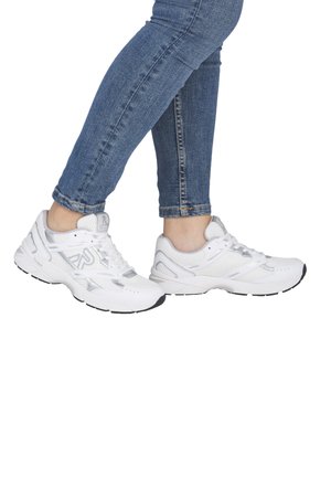 Person wearing blue jeans and white athletic shoes with silver accents, standing with one foot slightly raised on a white background.