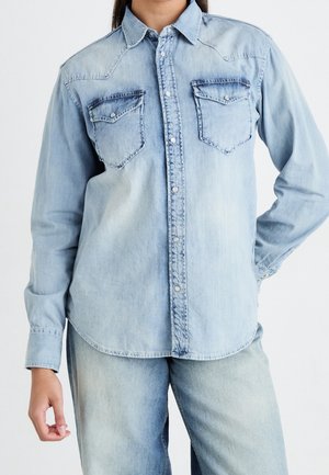 Mujer con camisa de mezclilla azul claro abotonada, con dos bolsillos en el pecho y jeans de mezclilla azul claro a juego, frente a un fondo liso.