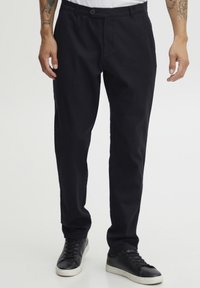 Mørk marineblå chinos laget av bomullsblanding, med knapplukking og sidelommer. Brukt med svarte joggesko, noe som fremhever en avslappet stil.