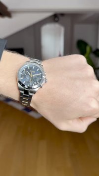 Relógio de pulso em aço inoxidável prateado com mostrador preto, detalhes em dourado e três submostradores. Tem uma bracelete metálica e exibição da data às 4 horas.