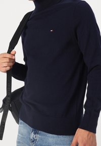 Mörk marinblå stickad tröja med mock neck, som har en liten logotyp på bröstet och ribbad mudd och nederkant, bärs med blå jeans och en svart väska.