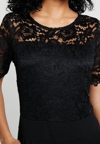 Top negro con un encaje superpuesto y mangas cortas, con un patrón floral texturizado. La sección inferior lisa contrasta con el encaje.