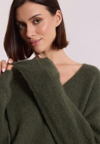 Zielony, oversize sweter wykonany z miękkiego, teksturowanego materiału, z głębokim dekoltem w serek i szerokimi rękawami; luźny krój podkreśla komfort.