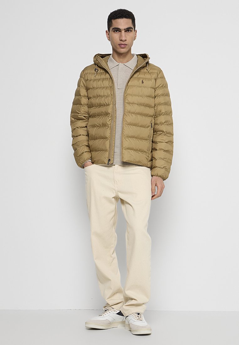 Beige Daunenjacke mit vertikaler Steppung, Kapuze und Reißverschluss. Getragen über einem hellgrauen Polohemd, kombiniert mit cremefarbenen Hosen und Sneakern.