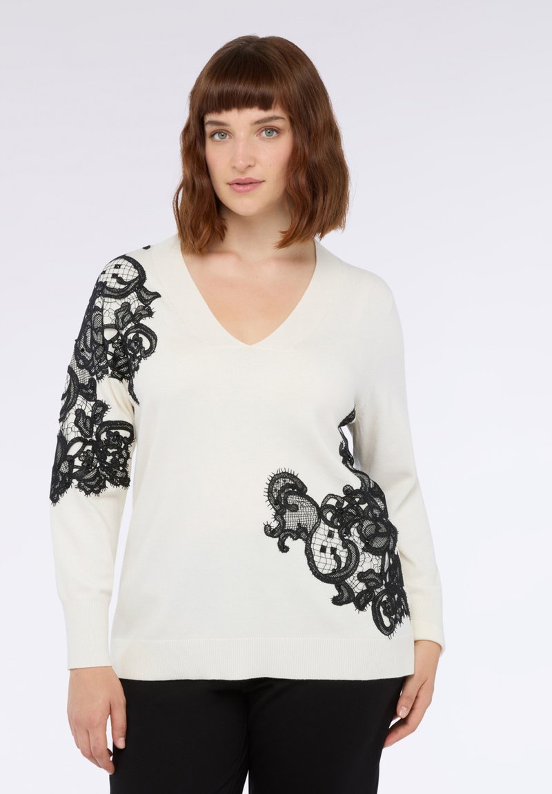 V-Ausschnitt Pullover in Creme mit schwarzen Spitzenakzenten am linken Ärmel und an der Seite, ausgestattet mit kunstvollen Mustern und einer lockeren Passform.