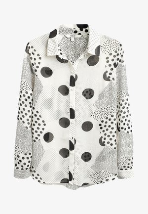 Femme portant un chemisier à pois, un pantalon en cuir noir, des talons hauts noirs à brides et une montre argentée, appuyée contre un mur blanc.