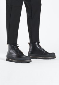 Stivaletti in pelle nera alla caviglia con punta rotonda, design con lacci e suola in gomma spessa con uno strato di sughero. Texture liscia con accenti minimi.