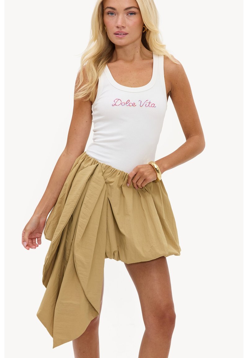 Femme blonde portant un débardeur blanc avec le texte "Dolce Vita" et une jupe beige courte avec un grand détail de tissu drapé et froncé.