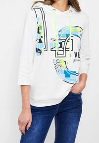 Sudadera blanca con gran estampado gráfico en azul y amarillo neón, presenta capucha, cordones y mangas de tres cuartos, combinada con jeans azules.