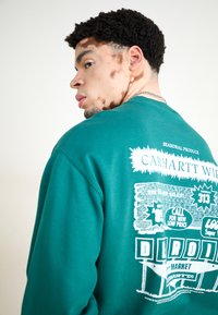 Turkusowy sweatshirt z graficznym nadrukiem na plecach, z białym tekstem i ilustracjami. Klasyczny fason z ściągaczem przy szyi i miękką strukturą.