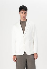 Blazer bianco su misura con due tasche frontali e bottoni beige, indossato sopra un dolcevita marrone, abbinato a pantaloni grigi. Design minimalista.