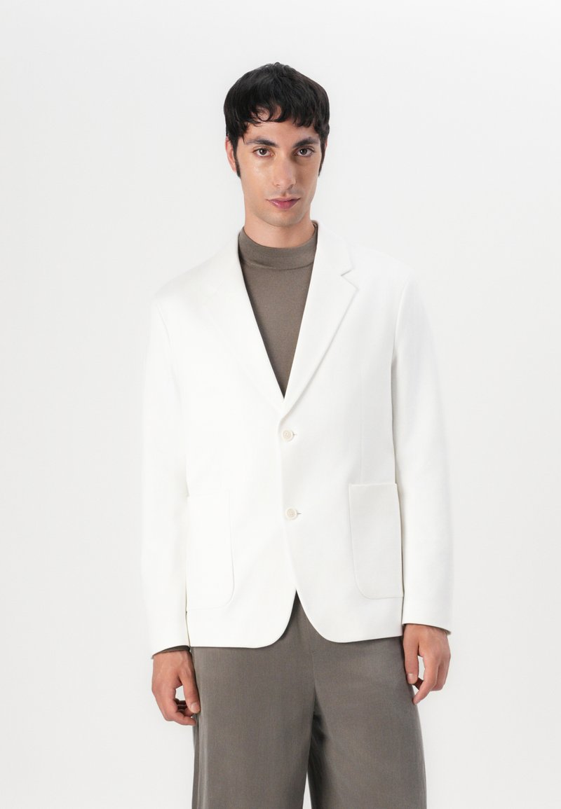 Blazer bianco su misura con due tasche frontali e bottoni beige, indossato sopra un dolcevita marrone, abbinato a pantaloni grigi. Design minimalista.
