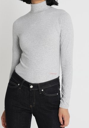 Maglione a collo alto grigio chiaro a maniche lunghe in tessuto morbido, caratterizzato da un piccolo logo rosso "Calvin Klein" e un design aderente. Abbinato a jeans di denim scuri.