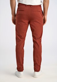 Pantaloni slim-fit color ruggine con una texture liscia, progettati con un frontale piatto, due tasche posteriori e orli risvoltati. Abbinati a sneakers bianche.