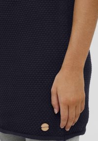 Marineblauer strukturierter Pullover mit geripptem Saum, ausgestattet mit einem Holzmarken-Tag in der unteren linken Ecke. Nahaufnahme einer Hand, die den Stoff greift.