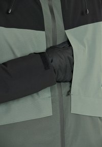 Sort og lysegrøn jakke med struktureret finish, lynlås foran, Velcro-detaljer og en lomme integreret i designet.