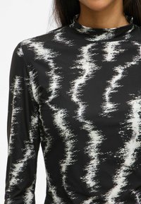 Femme portant un haut à manches longues noir avec un motif zigzag abstrait blanc et un col haut.