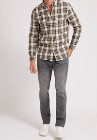 Geruit overhemd in groen, bruin en wit, met lange mouwen en twee borstzakken; gecombineerd met grijze jeans en witte sneakers.