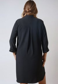 Zwarte jurk met lange mouwen, voorzien van een rugplooitje en knoopdetails op de manchetten. Glad textuur en een klassieke, gestroomlijnde vorm.