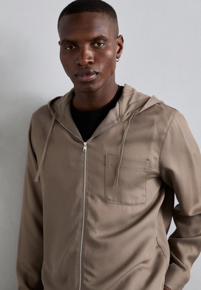 Φούτερ ζακέτα με κουκούλα σε μπεζ ύφασμα, με μπροστινή τσέπη, λεία υφή και κορδόνια στην κουκούλα.