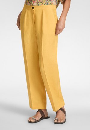 Persoon draagt mosterdgele wijde broek, bloemen top en donkerbruine sandalen met zilveren accenten, staand tegen een effen achtergrond.