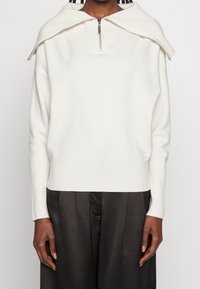 Witte gebreide trui met een brede, opgevouwen kraag en een halve ritssluiting; geribbelde manchetten en zoom; minimale logodetail aan de zijkant.
