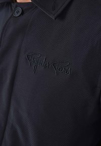 Schwarze Jacke aus strukturiertem Stoff, mit hohem Kragen, Knopfverschluss und gesticktem "Project x Paris" Logo auf der Brust.