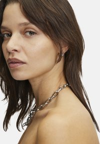 Sølv kæde med forlængede ledder, båret om halsen, suppleret af en lille hoopearring. Huden har en naturlig tekstur.