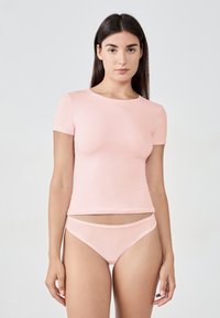 Eine Frau mit langen dunklen Haaren, die ein passendes, hellrosa kurzärmliges Oberteil und hochgeschnittene Bikinihöschen trägt, steht vor einem schlichten hellen Hintergrund.