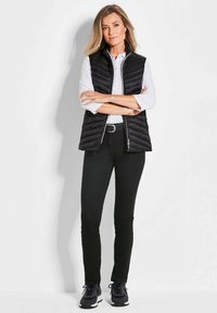 Schwarze Pufferweste mit Reißverschluss, getragen über einem weißen Hemd mit Kragen. Schwarze Slim-Fit-Jeans und dunkle Sportschuhe runden das Outfit ab.