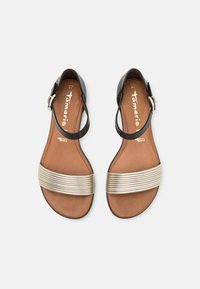 Platta sandaler i svart och guld, med ett texturerat guldband över foten och ett justerbart ankelsnöre. Brun innersula med logotyp.