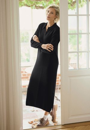 Nainen pitkässä mustassa mekossa ja ruskeissa kengissä seisoo käsivarret ristissä valkoisen oven vieressä, taustalla näkyy ulkopation kalusteita ja kasvillisuutta.