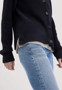 Persoon in lichtblauwe spijkerbroek en een zwarte cardigan met knopen over een beige shirt, gefotografeerd van de taille tot halverwege de dijen.