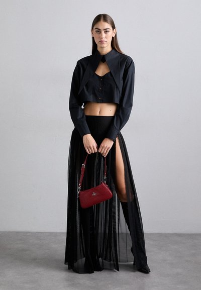 Μαύρο κοντό πουκάμισο με αιχμηρό γιακά και λεπτομέρειες κουμπιών, συνδυασμένο με μια διάφανη μαύρη φούστα και μπότες μέχρι το γόνατο σε μαύρο χρώμα. Κρατάει μια κόκκινη τσάντα ώμου.