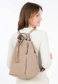 Beige perforerad ryggsäck med svart kantning och en lång tofsdetalj. Har justerbara remmar och en silverfärgad dragkedja för åtkomst till huvudfacket.