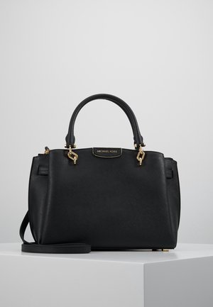 Černá kabelka Michael Kors se zlatým kováním, dvojitými rukojeťmi a odnímatelným ramenním popruhem, umístěná na bílém podstavci.