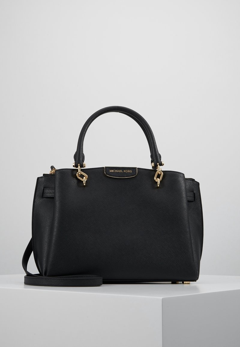 Czarna torebka Michael Kors ze złotymi okuciami, podwójnymi uchwytami i odpinanym paskiem na ramię, umieszczona na białym podeście.
