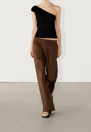 Femme portant un top côtelé noir à une épaule, un pantalon marron ample avec cordon de serrage, et des sandales noires en tongs, debout devant un fond uni.