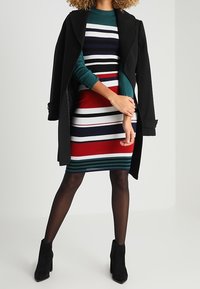 Randig klänning i röd, marinblå och grön, med långa ärmar och en åtsittande silhuett, matchad med en svart jacka och högklackade stövlar.