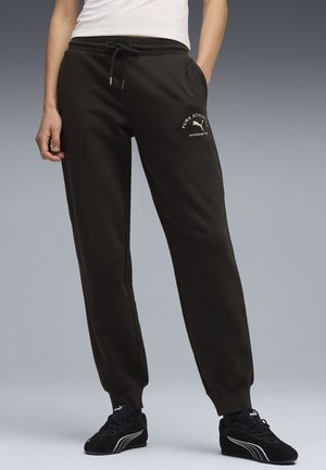 Persona con pantalones deportivos negros Puma con el logo en el muslo, zapatillas negras Puma y una camiseta blanca ajustada, de pie frente a un fondo gris.